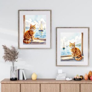 Puede incluir: Dos impresiones en acuarela enmarcadas representan gatos atigrados naranjas junto a una ventana con vistas a un paisaje marino con veleros. La obra de arte presenta un día soleado con agua y cielo azules, creando un sereno tema costero.