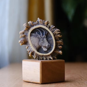 May include: A carved wooden plaque with a detailed image of a mountain goat's head. The plaque is mounted on a small wooden base.
