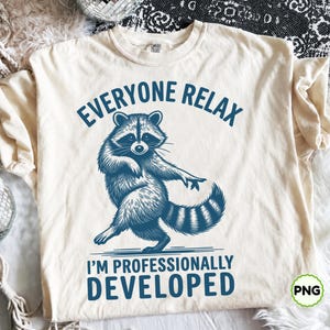 Puede incluir: Una camiseta de manga corta de color crema con un gráfico azul oscuro de un mapache de pie sobre sus patas traseras. El texto sobre el mapache dice EVERYONE RELAX y el texto debajo dice I'M PROFESSIONALLY DEVELOPED en una fuente azul negrita.