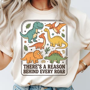Könnte beinhalten: Cremefarbenes T-Shirt mit einem rechteckigen Aufdruck, der verschiedene Cartoon-Dinosaurier in Grün-, Orange- und Gelbtönen zeigt. Der Aufdruck enthält den Text "THERE'S A REASON BEHIND EVERY ROAR" in schwarzer Schrift. Sterne und Blätter umrahmen das Design.