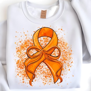 Pode incluir: Camisola branca com um grande design de fita de conscientização laranja. A fita, com contorno preto e laço, é colocada sobre salpicos de tinta laranja. Uma pequena etiqueta é visível perto da gola.
