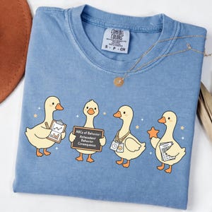 May include: Light blue Comfort Colors t-shirt featuring four cartoon ducks. Each duck holds an item: a clipboard, a chalkboard with text, a badge, and a star. The shirt has a round neckline and a small tag.