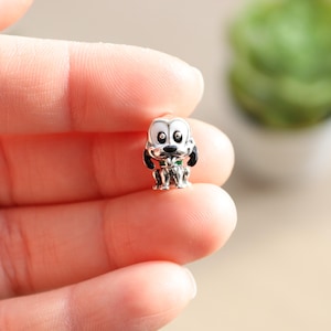 Könnte beinhalten: Silberner Anhänger mit einem Cartoon-Hund mit weißem Fell, schwarzen Flecken und grünem Halsband. Der Hund hat schwarze Augen und eine rosa Zunge, die herausragt.