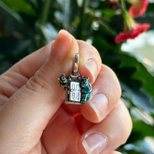 Puede incluir: Un charm de plata con un monstruo verde y amarillo y un oso verde de pie junto a una pequeña puerta blanca con marco negro. El charm tiene un lazo de plata en la parte superior para sujetarlo a una pulsera o collar.
