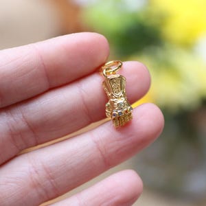 Pode incluir: Um pingente dourado em forma de manopla, com pedras preciosas embutidas em várias cores. O pingente é suspenso por um pequeno laço, sugerindo que foi projetado para um colar ou pulseira. Os detalhes da manopla são finamente trabalhados.