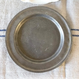 May include: A round, antique pewter plate with a slightly textured surface. The plate has a wide rim and a shallow center. The color is a muted gray with some darker spots, suggesting age and use. The plate is set on a linen tablecloth.