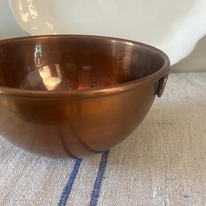 May include: A copper bowl with a rounded shape and two small handles. The bowl has a shiny, reflective surface and sits on a striped fabric. A white, scalloped-edge plate is in the background.