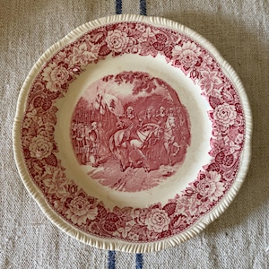 May include: A round ceramic plate with a cream-colored base and a deep red transferware pattern. The rim features a detailed floral border, while the center depicts a historical scene of soldiers on horseback carrying a flag with a pine tree.