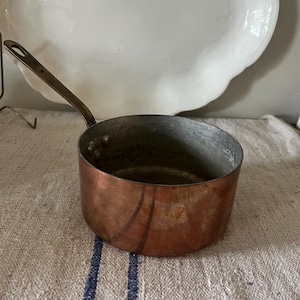 May include: A small copper saucepan with a long, brass handle. The pot has a rounded shape and a slightly tarnished appearance, suggesting age. A white, scalloped-edge serving dish is in the background. The pan sits on a striped, textured fabric.