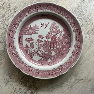 May include: A round, white plate with a pink, detailed Willow pattern design. The plate features a scalloped edge and a central scene with pagodas, trees, and birds. The border has a repeating pattern of geometric shapes and floral elements.