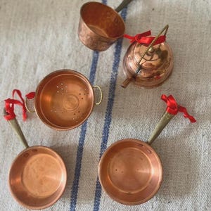 May include: A collection of miniature copper kitchenware items including two frying pans, a colander, and a kettle. Each piece features a metallic copper finish, brass-toned handles, and is adorned with small red ribbon bows tied to the handles.