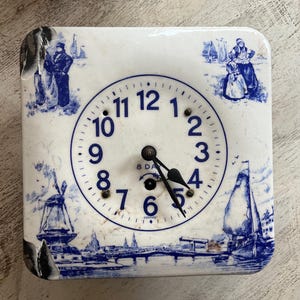 May include: Square, vintage clock with a white face and blue illustrations of Dutch scenes. The clock face has black hands and numbers. The clock has the text "8 DAYS GERMANY".