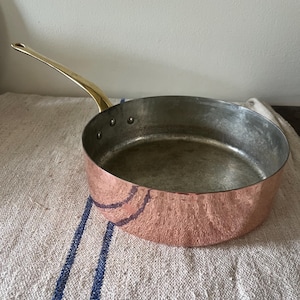 May include: A round copper sauté pan with a polished brass handle attached by three rivets. The exterior features a hammered texture, while the interior is lined with a silver-colored metal. The pan has a wide, flat base and straight sides.