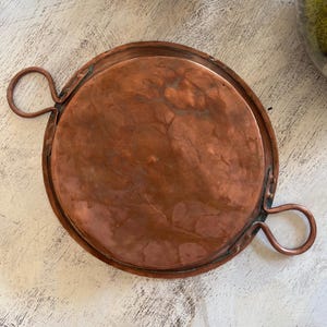 May include: A round copper tray with two loop handles. The tray has a slightly raised edge and a rich, reddish-brown color. The handles are attached to the sides of the tray and are also copper. The tray appears to be vintage or antique.