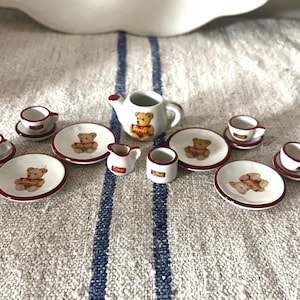 May include: A miniature Steiff teddy bear tea set. The set includes a teapot, cups, saucers, plates, a creamer, and a small mug. Each piece is white with a red trim and features a teddy bear illustration. The set is arranged on a white and blue striped surface.