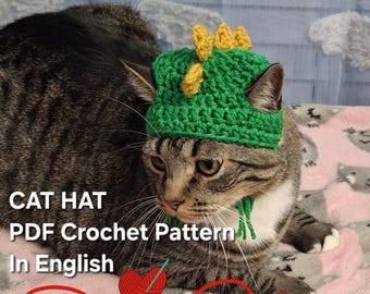 May include: A gray tabby cat wearing a green and yellow crocheted cat hat. The hat has a pointed top with yellow spikes. The cat is lying on a pink and gray blanket. The text "CAT HAT PDF Crochet Pattern In English" is visible in the image.
