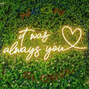 Puede incluir: Letrero de neón con la frase "It was always you" en cursiva, con un símbolo de corazón al final. El letrero se ilumina en un color amarillo cálido y se coloca sobre un fondo de follaje verde.