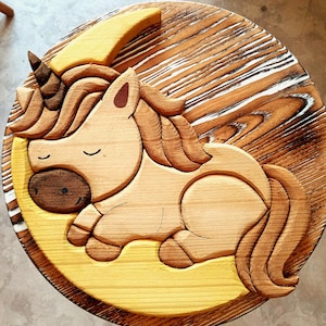 May include: A wooden carving of a unicorn sleeping on a crescent moon. The unicorn is light brown with a yellow horn and mane. The moon is yellow and the background is a wooden surface.