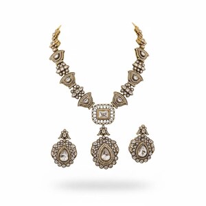 Può includere: Un set di collana e orecchini in metallo color oro antico con dettagli in filigrana. La collana presenta un ciondolo rettangolare centrale e una grande goccia, mentre gli orecchini sono pezzi coordinati a forma di goccia.