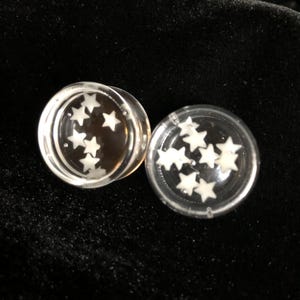 May include: Two clear, circular ear gauges with white star-shaped designs inside. The gauges are made of a transparent material and are set against a dark background. The stars are evenly spaced within the gauges.