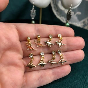 以下が含まれることがあります： 金色の恐竜のイヤリングのコレクション。 いくつかのイヤリングは小さなオレンジ色の恐竜のデザインが特徴で、他のイヤリングは上部に透明な丸い宝石が付いています。 イヤリングは手の中に表示されています。
