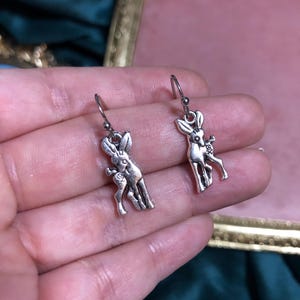以下が含まれることがあります： シルバーカラーの鹿のイヤリングのペア。各イヤリングは、シンプルなフックから吊り下げられた、花のアクセントが付いた詳細な鹿のペンダントが特徴です。イヤリングは、柔らかくぼやけた背景に対して表示されます。
