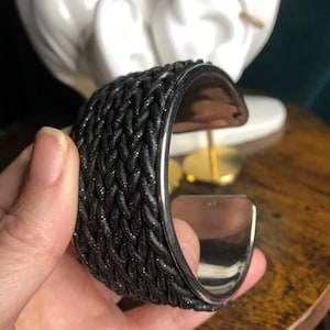 Könnte beinhalten: Ein schwarzes Armband mit geflochtenem Design und silberfarbenen Kanten. Das Armband hat ein strukturiertes Aussehen mit Goldflocken im gesamten schwarzen Material. Die Innenseite des Armbands ist silberfarben glänzend.
