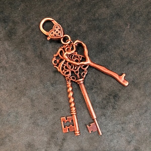 May include: A copper-colored keychain featuring three decorative keys. The keychain has a heart-shaped clasp and a key with a heart-shaped design. One key has a twisted handle, and another has a simple, straight design. The keys are detailed with intricate patterns.