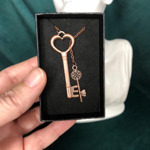 May include: A copper-colored key pendant necklace with a heart-shaped top and a smaller key charm. The necklace is displayed in a black box against a dark green background. The key pendant is a vintage-inspired accessory.