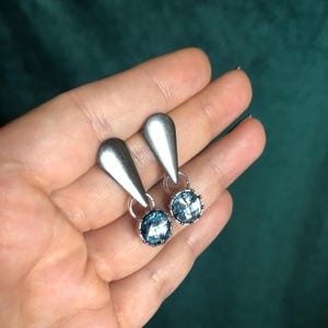 May include: A pair of silver-toned drop earrings. Each earring features a teardrop-shaped top and a round, light blue gemstone with a decorative silver setting. The earrings are displayed against a dark green background.