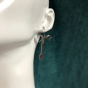May include: Silver-toned fork earrings with a bow detail. The earrings feature a small bow at the top, with a miniature fork charm hanging below. The earrings are designed to be worn with a standard ear piercing.