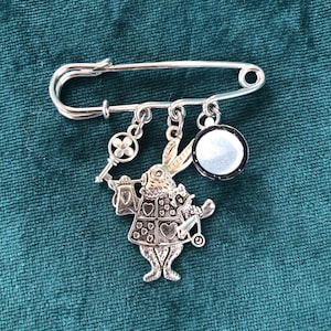 Broche de plata con dije: llave, conejo heraldo y taza de porcelana.