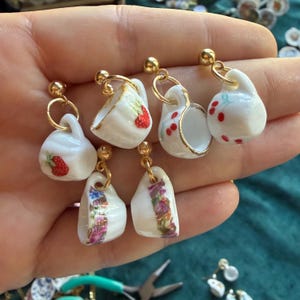 May include: Six miniature ceramic teacup charms featuring gold-tone metal loops. Two cups display red strawberry patterns, two feature red cherry motifs, and two are decorated with colorful floral bands. Each piece has a white glossy finish.