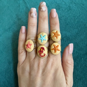Könnte beinhalten: Auf einer Hand sind sechs goldfarbene Ringe zu sehen, jeder mit einem cremefarbenen Oval mit einem Meereskreaturen-Design. Die Designs umfassen Seesterne in Rosa, Orange und Blau sowie ein rotes Seepferdchen. Der Hintergrund ist türkis.