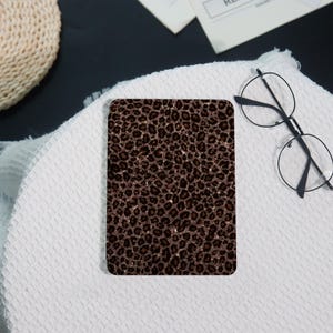 Peut inclure: Un objet rectangulaire avec un imprimé léopard marron et une texture scintillante. Il est posé sur une surface blanche texturée, avec des lunettes à monture ronde et un panier en osier en arrière-plan.