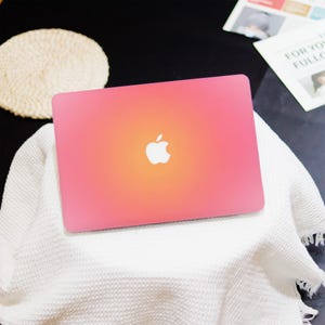 May include: A pink laptop with an Apple logo on the lid. The color gradient transitions from pink to orange in the center. The laptop is resting on a white, textured blanket.