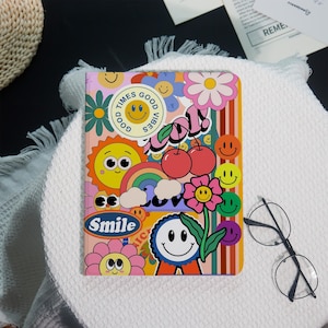 May include: A colorful notebook with a retro design featuring smiley faces, flowers, rainbows, and cherries. The notebook has the words "Times Good Vibes" and "Smile" printed on it. The cover is a mix of pink, orange, and green.