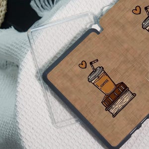 May include: A clear phone case with a brown, wood-grain design. The design features a coffee cup, a stack of books, and a heart. The word "COFFEE" is printed on the cup. The case is on a white, textured surface.
