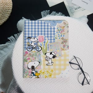 May include: A rectangular notebook cover featuring a collage of Snoopy and Woodstock illustrations. The design includes blue and yellow gingham patterns, floral motifs, and scenes of Snoopy riding a bicycle and holding a drink. The cover has a smooth finish.