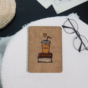 May include: A rectangular coaster with a brown background features a coffee cup illustration atop two books. The coffee cup has the word "COFFEE" printed on it, and a heart floats above it. A straw is in the cup. The coaster is on a white surface.