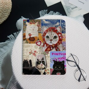 May include: A decorative notebook featuring a collage of cat illustrations. The artwork includes a white cat with strawberries, a cat in a red hood, and other cats with wine and coffee. The word "PORTUGAL" is visible on the cover.