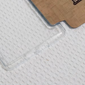 May include: A clear, transparent phone case with a protective bumper. The case is partially covering a wooden phone back with a design. The background is a white, textured surface.