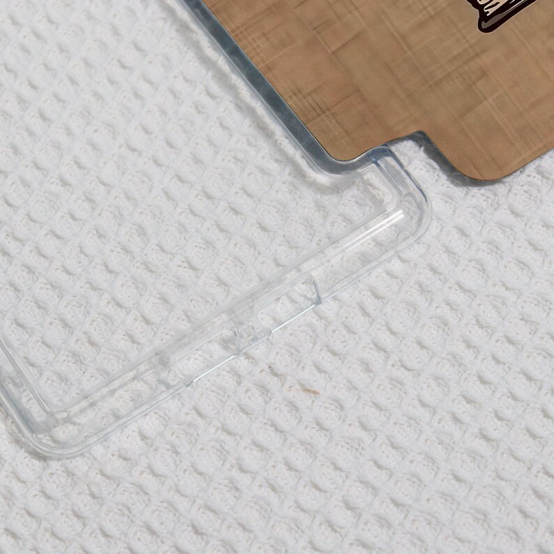 May include: A clear, transparent phone case with a protective bumper. The case is partially covering a wooden phone back with a design. The background is a white, textured surface.