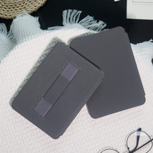 May include: Two gray tablet cases are displayed on a white textured surface. One case is open, revealing a gray elastic strap for support. The other case is closed. The cases are rectangular and appear to be made of a smooth material.