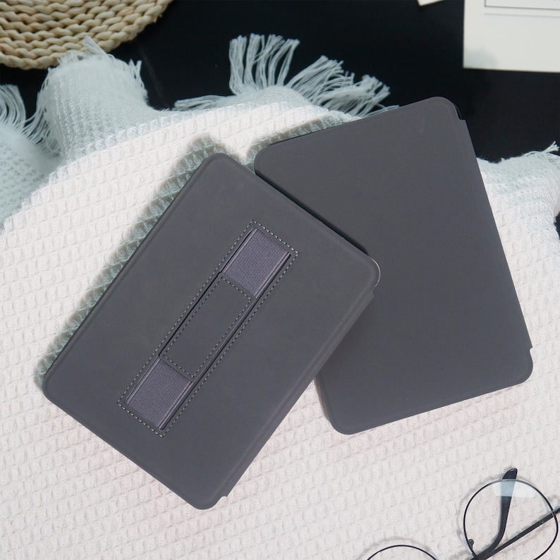 May include: Two gray tablet cases are displayed on a white textured surface. One case is open, revealing a gray elastic strap for support. The other case is closed. The cases are rectangular and appear to be made of a smooth material.