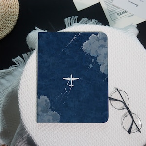 May include: A navy blue notebook with a white airplane illustration flying through clouds. The notebook is rectangular and has a minimalist design. The image also includes a pair of round eyeglasses.