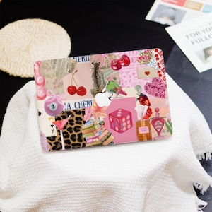 May include: A pink laptop case with a collage design featuring cherries, leopard print, dice, and perfume bottles. The case has a variety of images and text, creating a unique and artistic aesthetic.