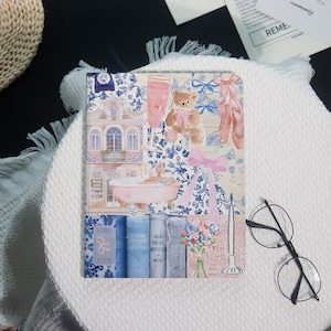 May include: A decorative journal with a collage design featuring a teddy bear, ballet shoes, and a woman in a bathtub. The background has a blue and white floral pattern. The journal is resting on a white surface.