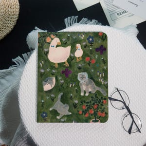 May include: A green notebook with a whimsical illustration of geese, cats, and flowers. The cover features a duck with a blue bow tie, a smaller duckling, and several cats. The design includes red, purple, and blue flowers.