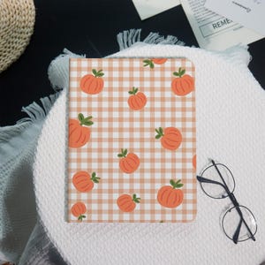 Könnte beinhalten: Ein Notizbuch mit einem pfirsichfarbenen Vichy-Muster und Illustrationen von orangefarbenen Kürbissen. Die Kürbisse haben grüne Stiele und sind über das Cover verteilt. Das Notizbuch liegt auf einer weißen, strukturierten Oberfläche.
